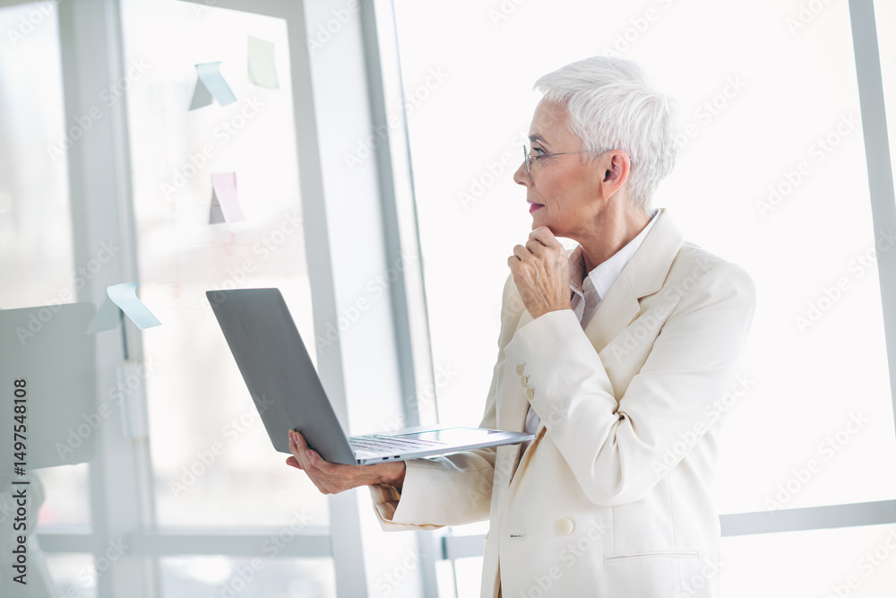 Confident senior businesswoman in elegant white suit analyzing strategy while using laptop in bright modern office Confident senior businesswoman in elegant white suit analyzing strategy while using laptop in bright modern office