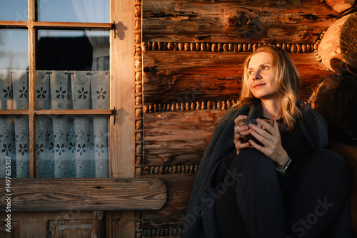 Woman wrapped in cozy blanket, savoring warm drink while sitting peacefully by window in rustic mountain chalet at sunset, enjoying tranquility of golden hour. Woman with tea in front of mountain hut photo