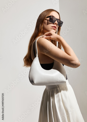 A stylish woman in sunglasses and with a white leather bag in her hand poses against a light background in fashionable summer clothes
