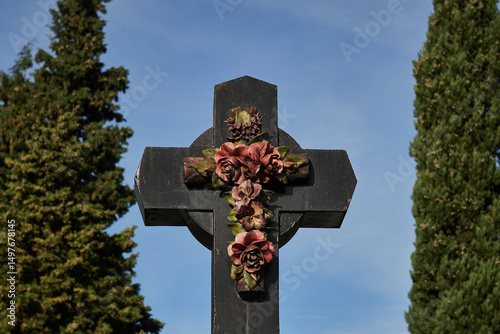 Photograph of a black stone funerary cross adorned with sculpted floral reliefs in shades of pink and green, set against a clear sky and flanked by tall cypress trees.
