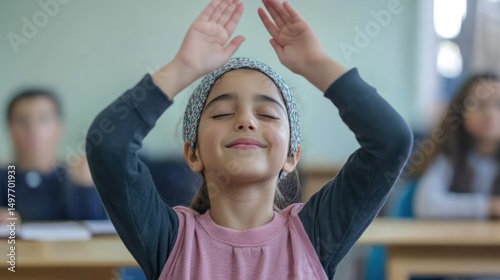 Girl meditating in classroom, eyes closed, hands above head.