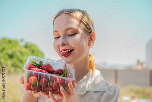 Young woman holding container with fresh strawberries and smiling, licking lips. Enjoyment of fruits. Plastic disposable packaging with berry for food in human hands