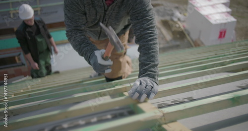 Worker using hammer to secure nails on roof frame, wearing gloves and helmet, close-up view of hands and tool on construction site, part of building project teamwork