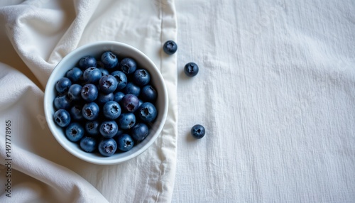 a top down view of a white ceramic bowl filled with fresh blueberries placed on a light linen tablec