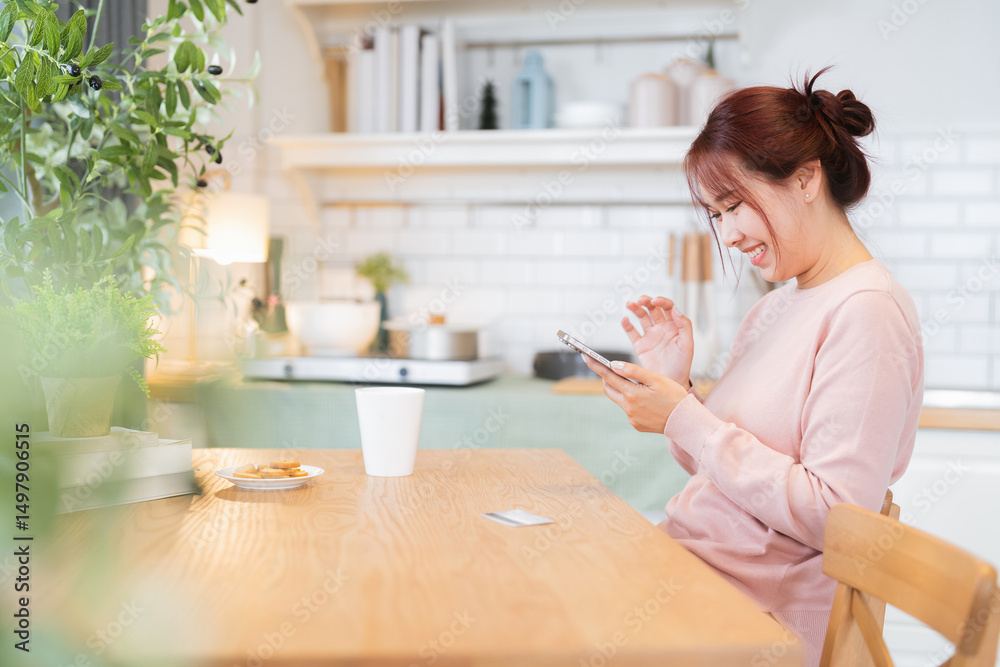 A beautiful young Asian woman smiles as she enjoys a cup of coffee in her cozy kitchen, using a smartphone and browsing a mobile app while relaxing at home A beautiful young Asian woman smiles as she enjoys a cup of coffee in her cozy kitchen, using a smartphone and browsing a mobile app while relaxing at home