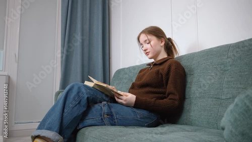 Student girl teenager comfortably rested reading a book, sitting on a soft sofa in a warm living room, enjoying a quiet moment of personal relaxation and literary research