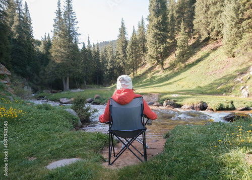 A girl or woman in a red jacket and white hat sits on a tourist chair and admires a mountain summer or spring landscape with a spruce forest, a blue river and yellow flowers in the morning. 