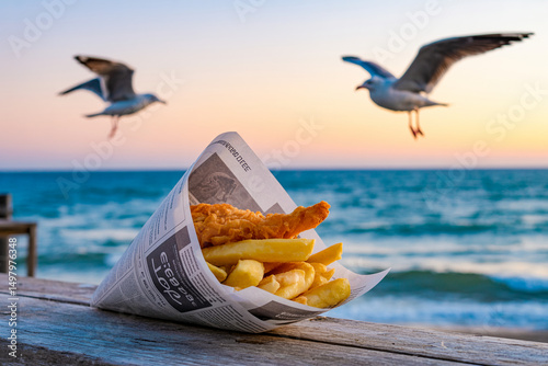 Crispy fish and chips in newspaper cone on rustic wood by ocean at sunset with seagulls, warm golden lighting and nostalgic seaside mood

