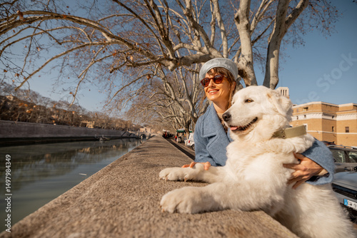 A joyful moment by the Tiber River in Rome as a woman and her fluffy white dog share the view and winter sunshine under bare sycamore branches, embracing the calm beauty of the Eternal City