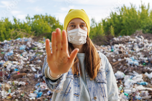 Принти на полотні A young woman in a yellow hat and a face mask holds up her hand in front of a landfill, conveying a message to stop pollution
