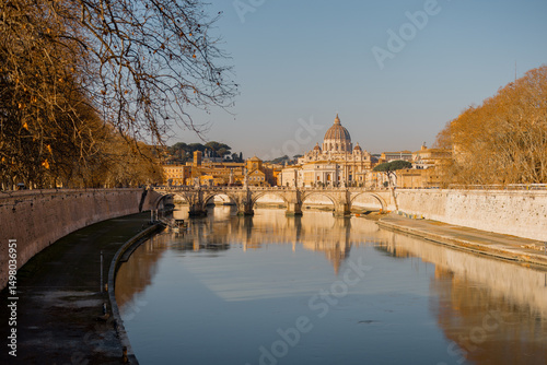 Fototapeta Naklejka Na Ścianę i Meble -  A peaceful morning view of St. Peters Basilica reflecting in the calm waters of the Tiber River, framed by winter trees and Roman bridges