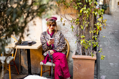Canvas Print A fashionable woman in a leopard print jacket and pink outfit sits outside a Roman cafe looking at her phone