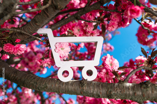 White shopping cart icon placed among blooming cherry blossoms on a tree with bright pink flowers and blue sky.