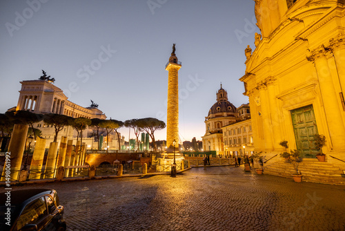 Fototapeta Naklejka Na Ścianę i Meble -  Evening view of Romes iconic Trajans Column and Altare della Patria, illuminated against the twilight sky, surrounded by ruins, churches and cobblestone streets