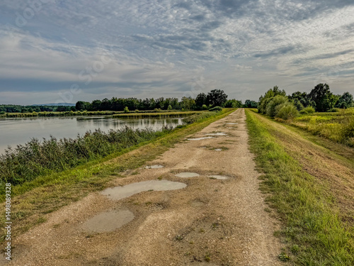 Fototapeta A gravel road with puddles, led along a flood embankment between the river and t