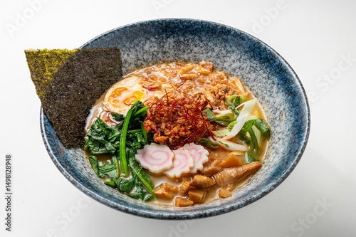 Spicy miso ramen with noodles, seaweed, spinach, egg, chili, narutomaki, and green onions in a rustic blue bowl on a white background.