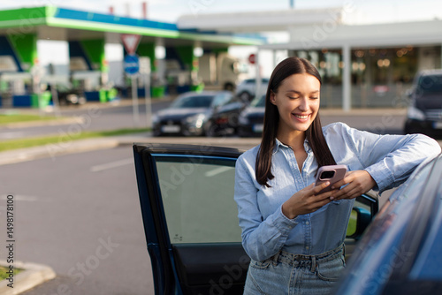 Woman driver resting after long driving, standing near her car and using cellphone, making stop at gas station, free space