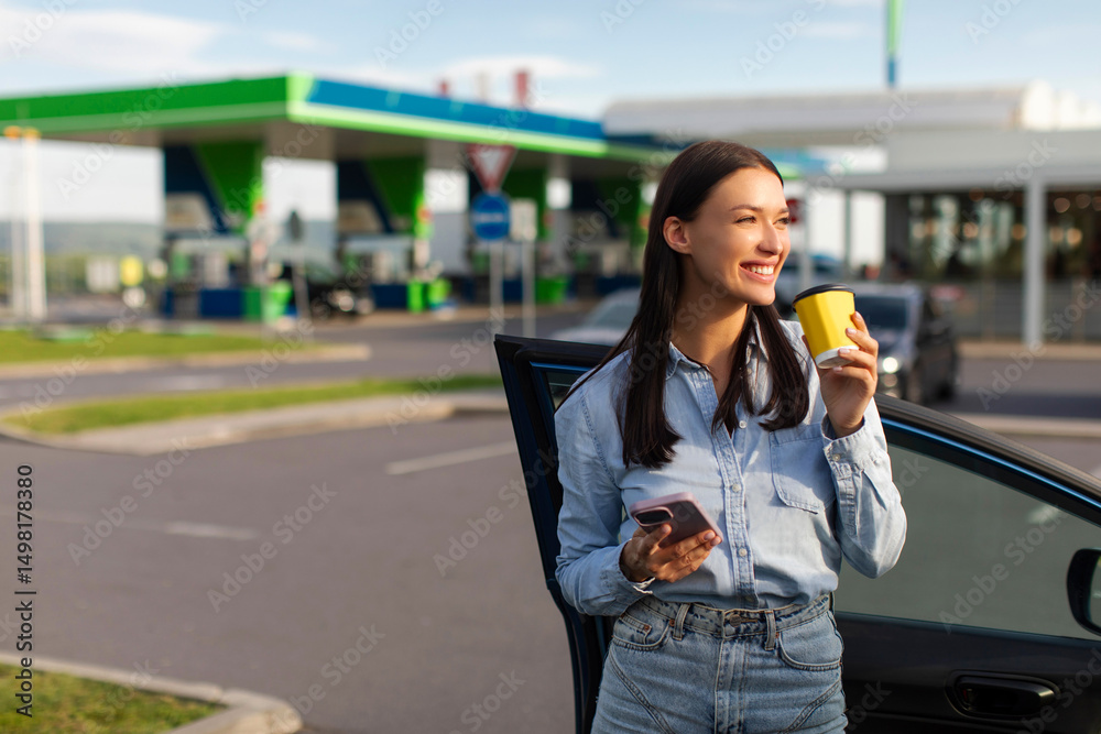Fototapeta premium Happy lady holding cellphone and drinking coffee, standing near her car on the petrol station parking, having break during road trip, copy space