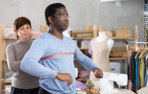 Woman tailor takes measurements from African American man client, using tape measure, measures body size for custom-made dress..