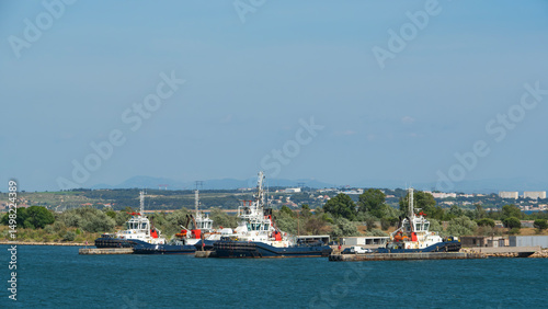 Four tugboats moored in a clear blue harbor, with green trees and a town visible on the distant shore under a bright sky