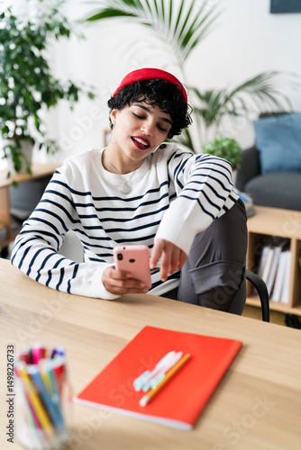 A young woman in a red beanie and striped shirt is using her phone at a desk. She's likely taking a break from work or studies, enjoying some downtime with her device.