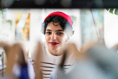 A woman in a red beanie smiles at someone through a glass barrier. She's likely interacting with someone while maintaining distance for safety.