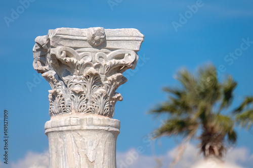 Corinthian capital of Kourion archaeological site in Cyprus with blurred palm tree and blue sky