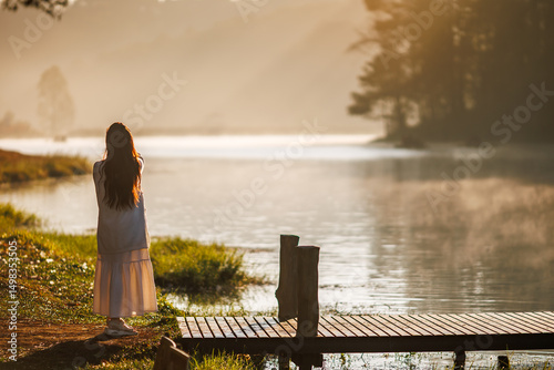 young woman stands on a serene wooden pier, gazing at a misty lake during a beautiful sunrise, with golden light reflecting on the water.
