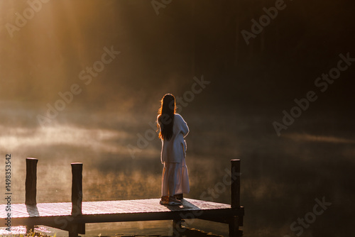 young woman stands on a serene wooden pier, gazing at a misty lake during a beautiful sunrise, with golden light reflecting on the water.