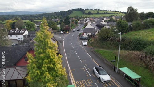 traffic on road passing by houses in village. Descending shot of moving vehicles and revealing bus stop. Wales, UK