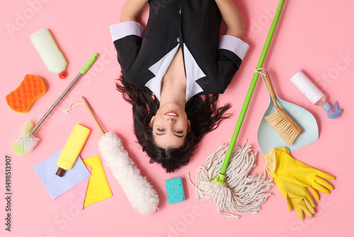 Happy young housemaid with different cleaning supplies lying on pink background