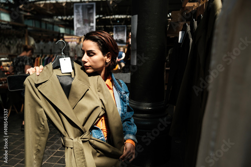 Young woman choosing trench coat in second hand clothing store photo