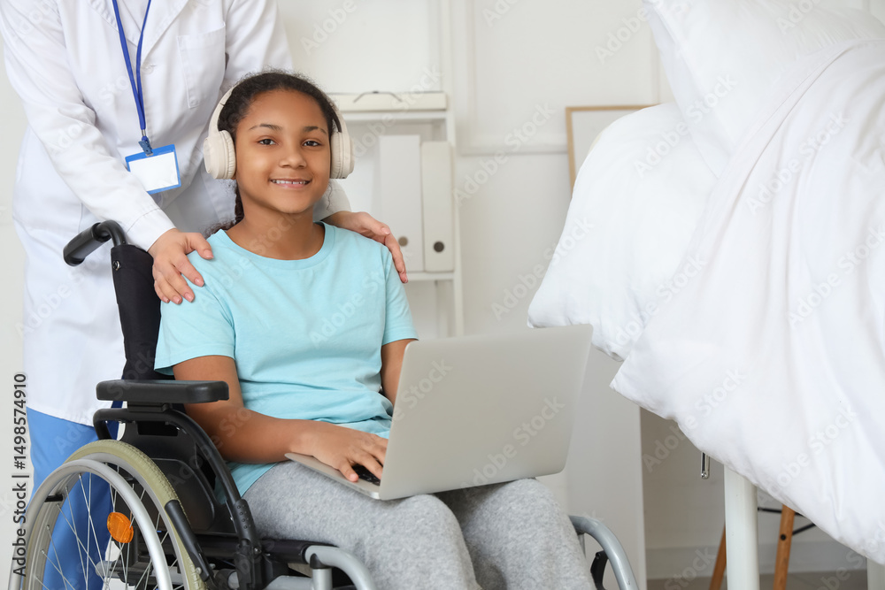 Obraz premium Smiling african american girl in wheelchair with headphones using laptop, doctor assisting in hospital room