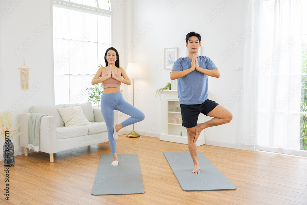 Obraz premium Asian man and woman practicing yoga together in bright living room, standing in tree pose on yoga mats, promoting mindfulness and balance in serene environment