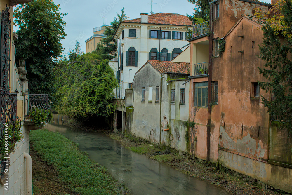 Fototapeta premium This serene scene captures a quiet canal flowing through the historic city of Padua, Italy. Lush greenery drapes over the banks, contrasting with the aged buildings lining the waterway.