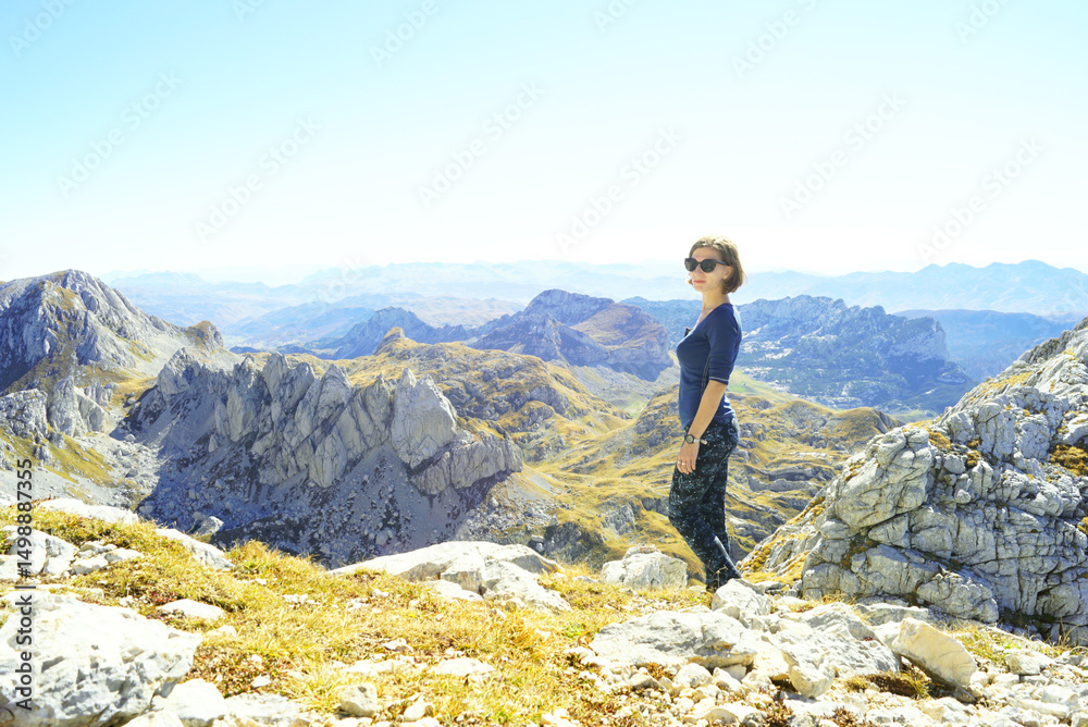 Fototapeta premium Hiking in the most beautiful places of Montenegro: a young smiling woman stands on the trail under the peak of Bobotov Kuk. Eco-tourism in Zabljak: a female tourist in the Durmitor National Park