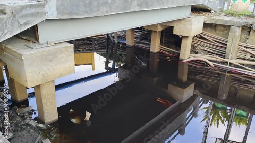 Urban drainage system with visible water, exposed utility cables, and concrete bridge supports.