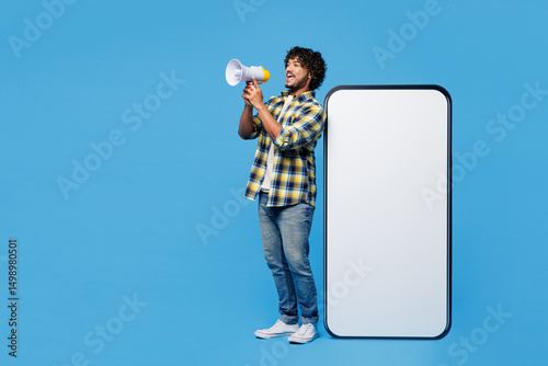 Full body young happy Indian man he wear yellow checkered shirt casual clothes big huge blank screen mobile cell phone smartphone with area scream in megaphone isolated on plain blue cyan background.