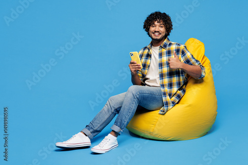Full body young Indian man he wears yellow checkered shirt casual clothes sit in bag chair hold in hand use mobile cell phone show thumb up isolated on plain blue cyan background. Lifestyle concept.