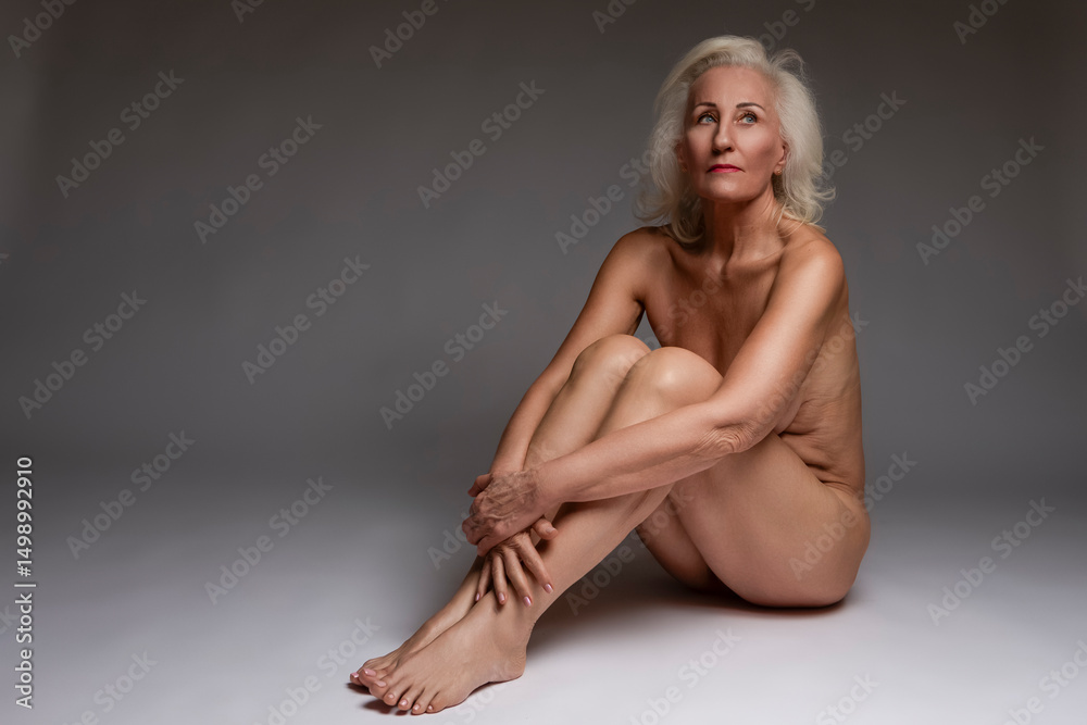 Fototapeta premium Senior Female Beauty Concepts. Portrait of Senior Nude Blond Caucasian Woman Sitting With Hands Crossed In Front Looking Upwards Posing On White