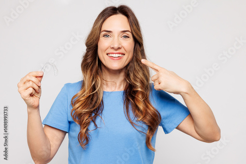 Young smiling woman wears blue t-shirt casual clothes hold in hand invisible transparent aligners, invisalign dental braces point on mouth isolated on plain white color background. Lifestyle concept.