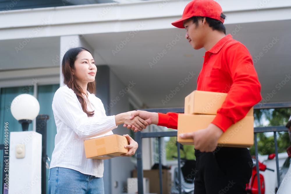 Obraz premium A delivery man, who appears to be Asian, in a red uniform shakes hands with a smiling woman, who seems to be middle-aged and holding a package, at the entrance of a modern home.