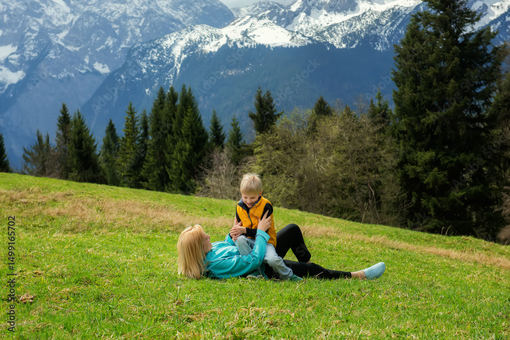 Naklejka premium Grandmother lying on green grass, playing with her happy grandson in yellow vest, surrounded by alpine mountains, trees, and nature. Joyful spring family moment outdoors.