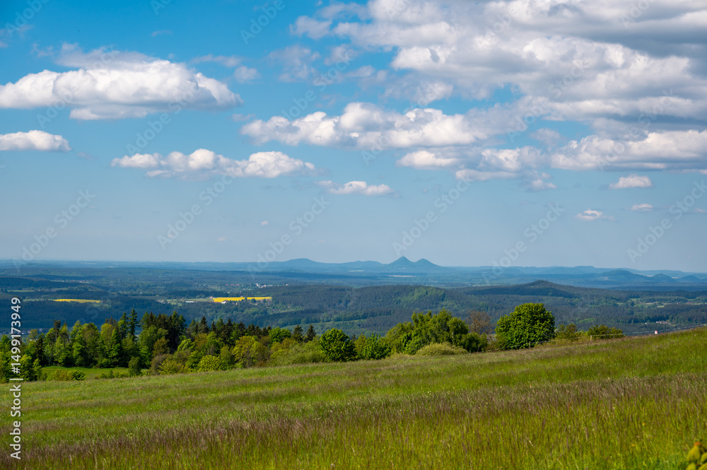 Fototapeta premium Panoramic view of green fields and forested hills near Jested, with the sharp peak of Ralsko mountain visible in the distance under a blue sky with scattered clouds.