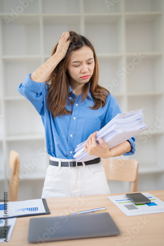 Young Asian office worker feeling stressed and tired while holding a pile of paperwork, showing frustration and exhaustion from overwork, inefficient work systems, and burnout photo