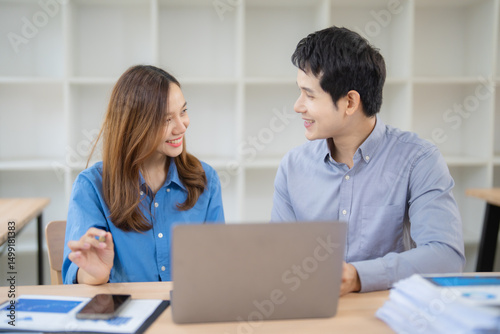 Two asian businesspeople are discussing and working together using a laptop computer, smiling and collaborating effectively in a modern office environment