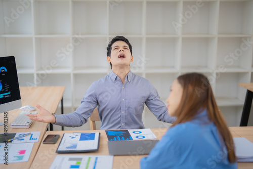 Stressed asian businessman complaining to his colleague about bad financial results, sitting at desk in office with computer and documents with charts and graphs