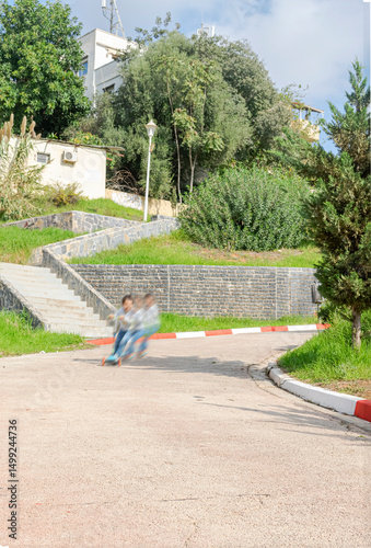 A child rides a scooter at high speed on a sunny suburban street park, creating a motion blur effect with greenery bushes and trees, stone walls, blue sky and residential buildings in the background.
