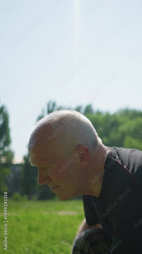 An elderly man squats down with a focused expression, seemingly engaged in a task, as sunlight reflects off him, the blurred background features a goalpost and lush greenery