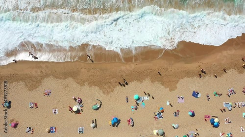 Overhead drone shot of anonymous tourist and colorful parasols on sandy seashore, showing people enjoying summer vacations at beach during sunny day